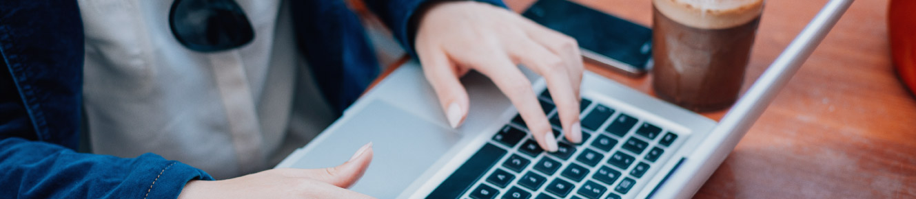 photo of individual typing on laptop with mobile device and cup of coffee beside them 